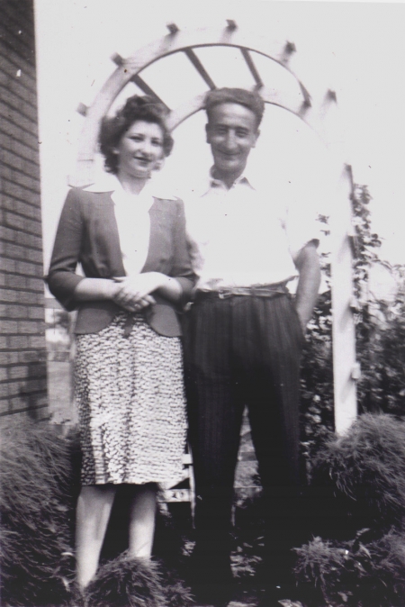 Photo en noir et blanc d'un homme et une femme se tenant ensemble à l'extérieur sous une tonnelle, souriant à la caméra. L'homme a les mains dans les poches.