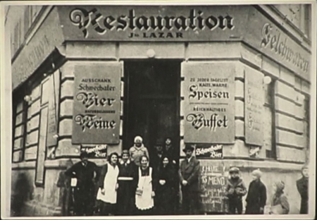 Photo en noir et blanc d'un groupe d'environ huit personnes se tenant devant l'entrée d'un restaurant. L'édifice a une grande affiche de chaque côté de la porte, rédigées en allemand. Deux jeunes enfants regardent le groupe d'un des deux côtés.