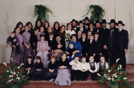 Colour photograph of a large group of about 30 people posing together for a photograph in 3 or 4 rows, some sitting in the front rows. Everyone is formally dressed. The women wear dresses and the men are in suits and hats. There are large bouquets of flowers on either side of the group.