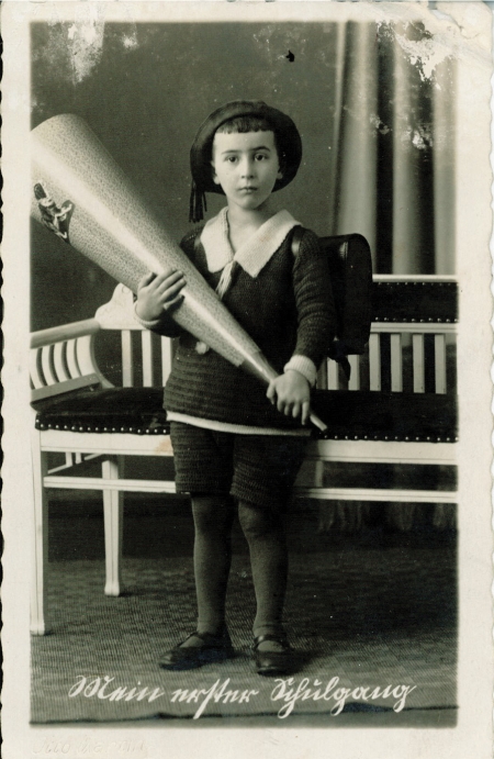 Portrait photographique pris en studio d'un jeune garçon en uniforme et chapeau d'écolier, regardant la caméra en adoptant une pose pensive. Il tient un gros objet ayant la forme d'un cornet. Il y a de l'écriture en allemand au bas de l'image, et les bords de la photo sont usés.