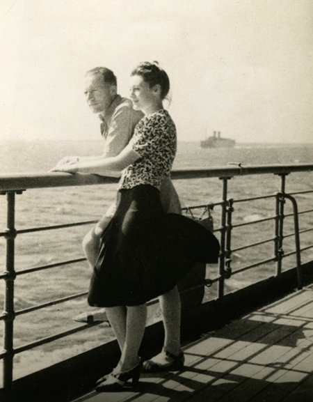 Black-and-white photograph of a woman standing beside a man on the deck of a ship. They both look ahead of them, leaning on the railing of the deck. There is water below them and a ship on the far horizon.