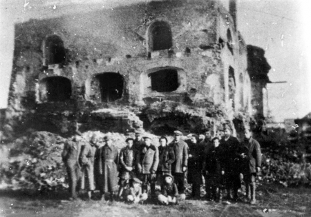 Black-and-white grainy photograph of a group of 15 people posing together outdoors in a line, 2 children sitting on the ground, in front of the ruins of a building. The building is missing a roof and has several window frames that no longer have any windows.