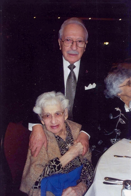 Photo en couleur de deux personnes âgées souriant à la caméra. L'homme est debout derrière la femme, assise sur une chaise, et ses mains sont sur ses épaules. Elle tient l'une de ses mains.