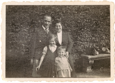 Photo en noir et blanc d'une femme et d'un homme debout ensemble avec deux jeunes enfants à l'extérieur, devant de grandes haies qui prennent tout l'espace en arrière-plan.