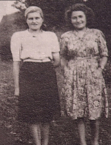 Black-and-white photograph of two young women standing and smiling outdoors. One woman wears a dress, and the other wears a blouse and skirt.