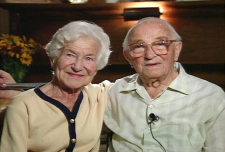 Colour photograph of an elderly couple sitting arm-in-arm and smiling at the camera. The woman on the left has short hair and wears a cardigan, and the man wears glasses and a collared shirt.