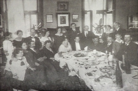 Black-and-white photograph of a group of about 20 people sitting and standing around a large dining table set with dishes. The group is formally dressed, and a couple sitting at the table appear to be celebrating their wedding.