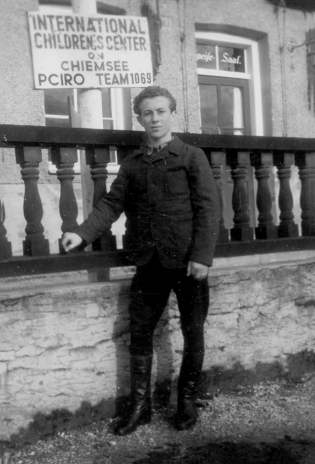 Black-and-white photograph of a young man standing outdoors on the grass and leaning against a raised  wooden railing behind him. A sign in the background reads ‘INTERNATIONAL CHILDRENS CENTER’.