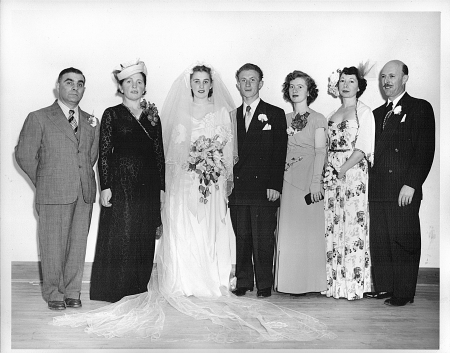 Photo en noir et blanc d'un groupe de sept personnes, se tenant en ligne et souriant à la caméra. Il semble que le couple au centre célèbre leur mariage puisque la femme est vêtue d'une longue robe blanche, d'un voile et tient un bouquet de fleurs. Les hommes du groupe portent des complets et les trois autres femmes des robes.