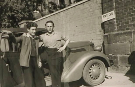 Black-and-white photograph of two young men standing together in front of a vintage car outdoors. One man has his arm around the other. A large brick wall is in the background.