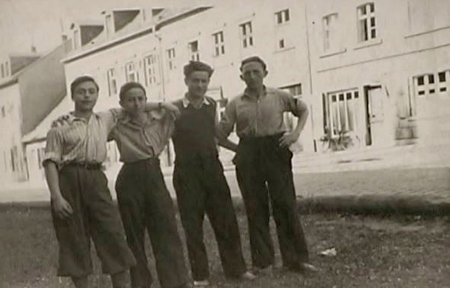 Black-and-white photograph of a group of four boys standing together arm-in-arm outdoors, in a shaded area, on a road. Across the street is a large building.