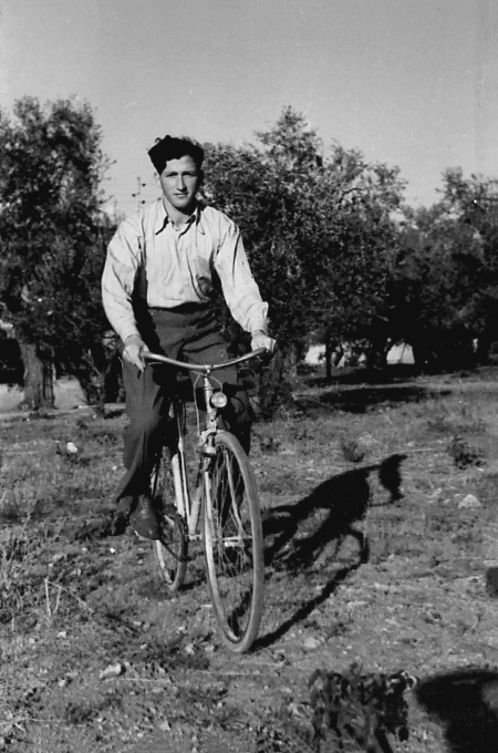 Photo en noir et blanc d'un jeune homme roulant à bicyclette à travers un champ. Il porte des pantalons foncés et une chemise à col de couleur pâle. Il y a des arbres en arrière-plan.