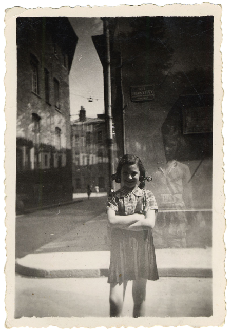 Black-and-white photograph of a young girl standing a street with her arms crossed, smiling at the camera. She stands near the corner of the street, with the road and sidewalk continuing in the background.