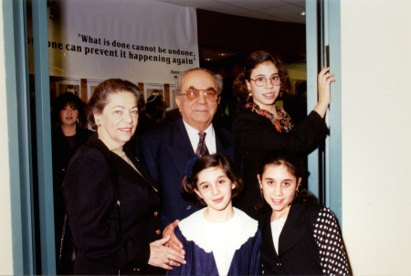 Colour photograph of an elderly man and woman, standing together indoors with a woman and two young girls. The family is formally dressed, with the girls wearing blouses and the man in a suit.