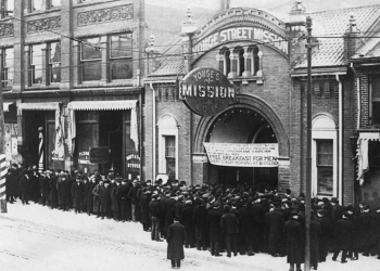 Black-and-white photograph of an outdoor street scene. A group of people are lined up outside a building.