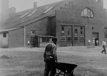 Black-and-white photograph of a man, with his back to the camera, pushing a wheelbarrow in a yard. There is a bench in the foreground, and a building in the far background. The man wears a uniform with a big circle on his back.