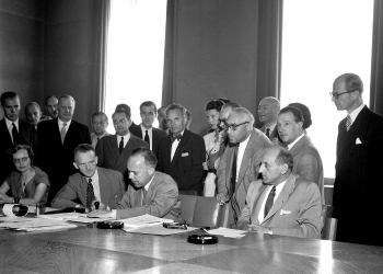 Black-and-white photograph of a group of about 25 people gathered together behind a large table in a conference room. Four men sit at the table, and one man is signing a document. The other people stand behind.