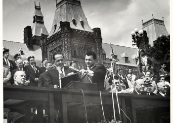 Photo en noir et blanc d'un homme en costume parlant dans deux microphones à un podium à l'extérieur. Il est entouré d'un groupe de gens, certains sont assis, un grand édifice est à l'arrière-plan.