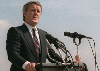 Colour photograph of a man in a suit, speaking outdoors at a podium hooked up with two microphones.