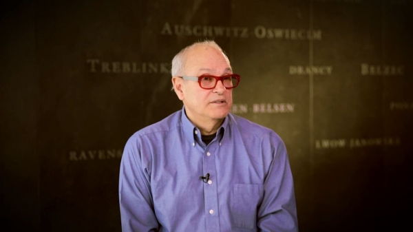 Screenshot from Harold Troper interview. He is sitting in front of a grey wall with various camps names written on it. His face and shoulders are visible at the camera.