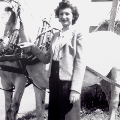 Photo en noir et blanc d'une femme aux cheveux bruns foncés, se tenant à l'extérieur auprès de deux chevaux blanc attachés à une voiture ou un charriot. Elle tient les rênes des cheveux d'une main.