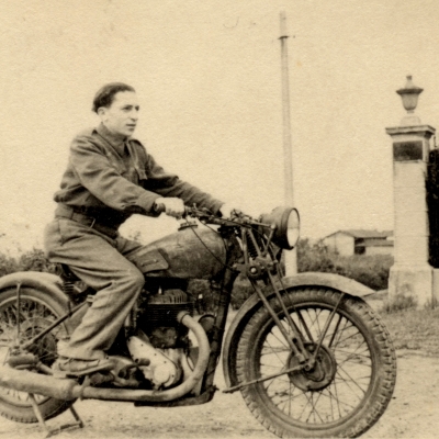 Photo en noir et blanc d'un jeune homme sur une motocyclette, regardant vers la droite de la caméra. Il y a une haie derrière lui et un bâtiment en arrière-plan.