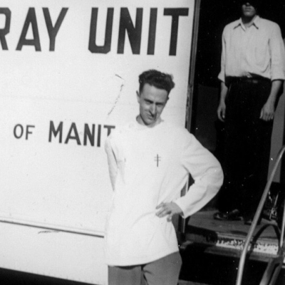 Photo en noir et blanc d'un homme debout, la main appuyée sur la hanche gauche, devant un grand camion blanc sur lequel il est inscrit “X-RAY UNIT” sur un côté. Un homme est debout derrière lu dans l'entrée du camion, son visage est caché par un ombrage.