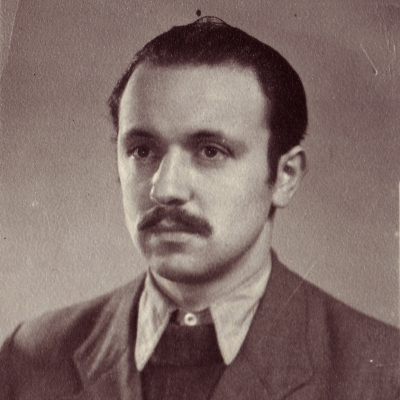 Sepia-toned portrait photograph of a man, pictured from the chest up, looking towards the left of the camera. He wears a suit and tie, and his hair is combed back. He has a moustache.