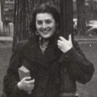 Black-and-white photograph of a woman smiling and leaning against a street light in front of a large field with buildings and cars in the background. She holds a purse and book in her hand.