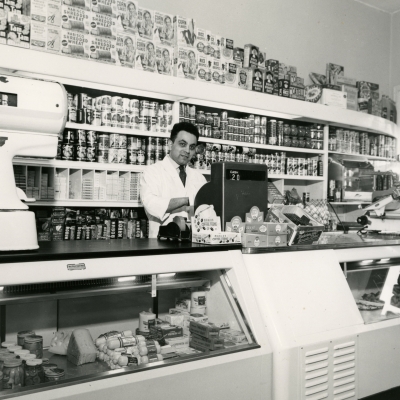 Black-and-white photograph of a man standing behind the cash register of  a large grocery store. He stands in front of a shelf full of food products, and there is a large display food counter in front of him.