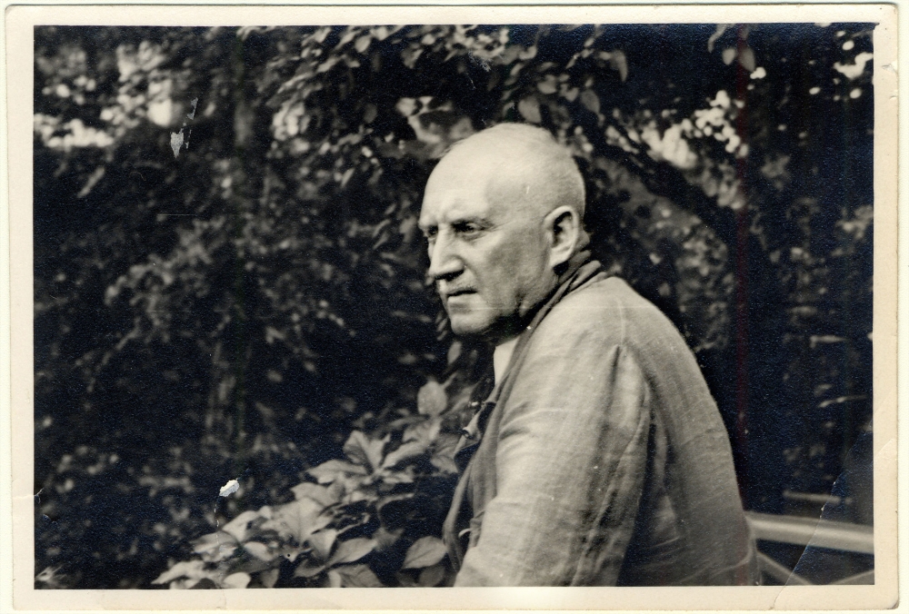 Black-and-white photograph of the side profile of an elderly man standing outdoors, surrounded by leaves and foliage. He looks away in the distance.