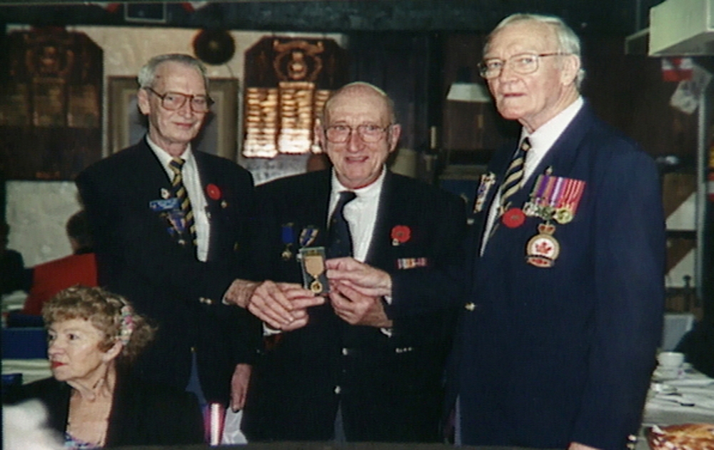 Photo en couleur de trois hommes âgés se tenant debout et tenant  une médaille militaire. Les hommes portent des vestons décorés de médailles et d'épingles.