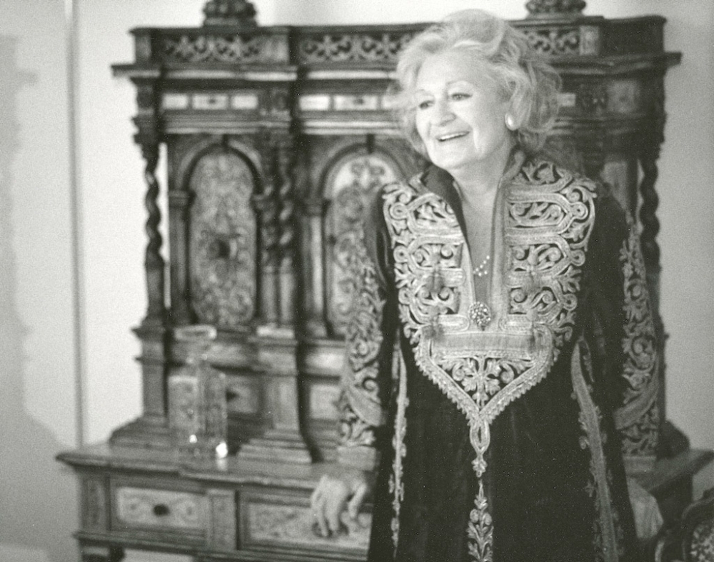 Black-and-white photograph of an elderly woman dressed in an embroidered tunic, leaning against an elaborately carved wooden hutch.