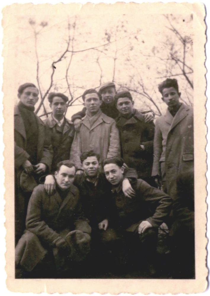 Photo en noir et blanc d'un groupe de neuf jeunes hommes se tenant ensemble bras-dessus bras-dessous. Ils portent des manteaux et il y a des arbres nus au-dessus d'eux en arrière-plan.