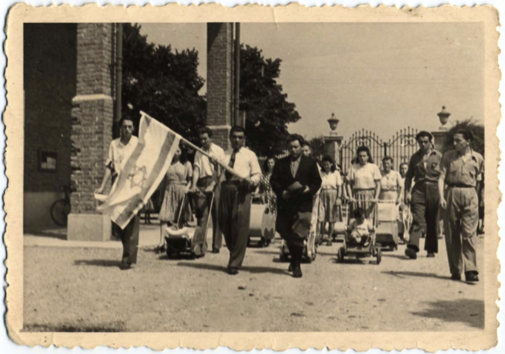 Black-and-white photograph of a parade or march with a large group people walking towards the camera. A man at the front of the crowd carries the flag of Israel, and several women in the background push strollers.