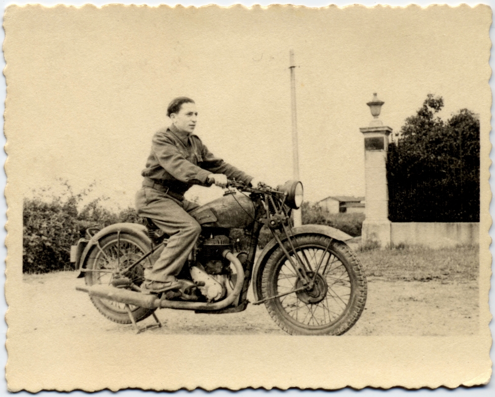 Black-and-white photograph of a young man posing on the back of a motorcycle, looking forwards towards the right of the camera. There is a hedge behind him and a building in the distant background.