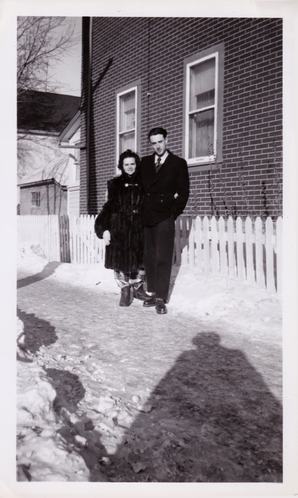 Black-and-white photograph of a woman and man standing together outdoors on a snowy driveway in front of a white fence. The woma