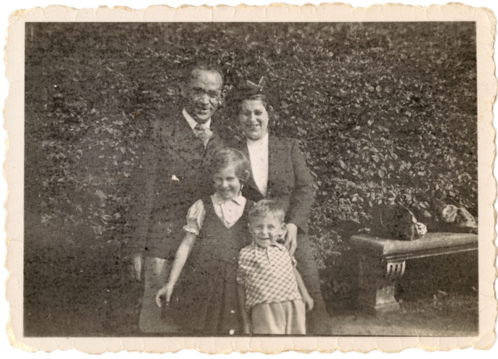 Black-and-white photograph of a woman and man standing together with two young children outdoors, in front of a large hedge that takes up the entire background.