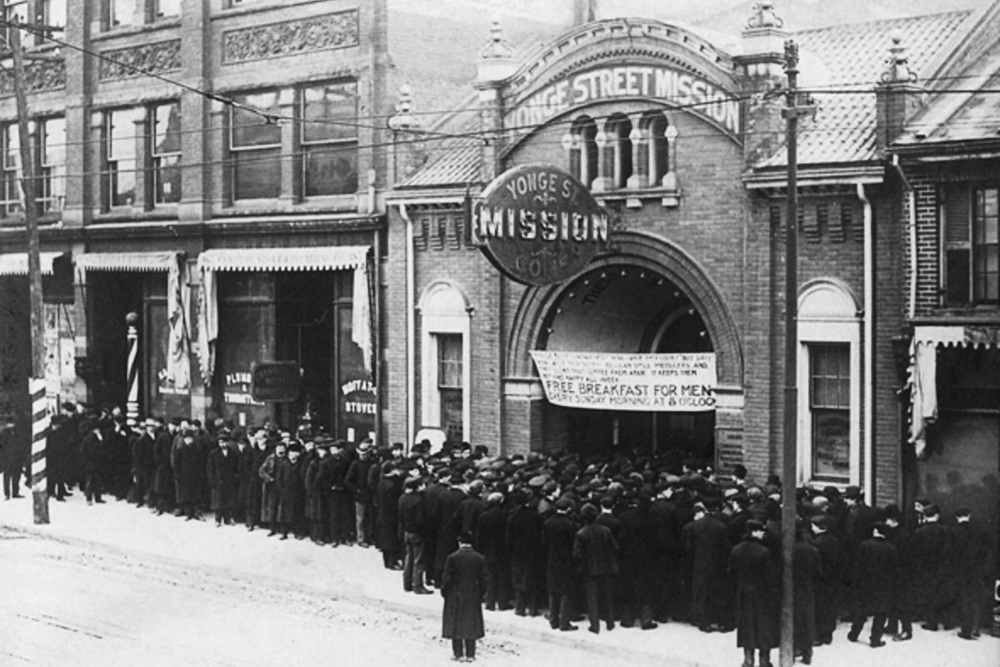 Black-and-white photograph of an outdoor street scene. A group of people are lined up outside a building.