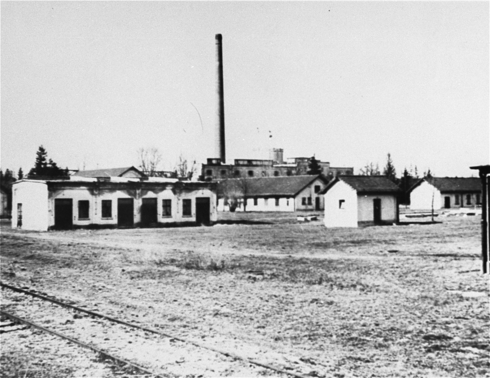 Photo en noir et blanc d'un champ avec des baraquements à un étage et une usine en arrière-plan.