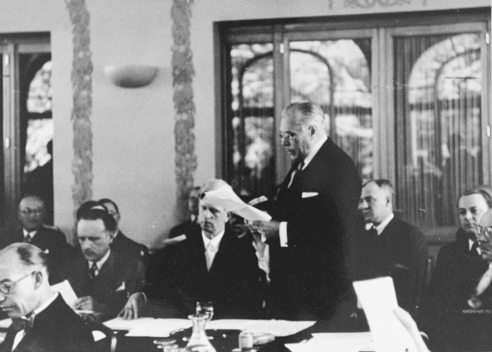 Black-and-white photograph of a man standing among a room of men who are seated at desks. The man standing up reads aloud from the paper he is holding, while wearing glasses and a business suit.