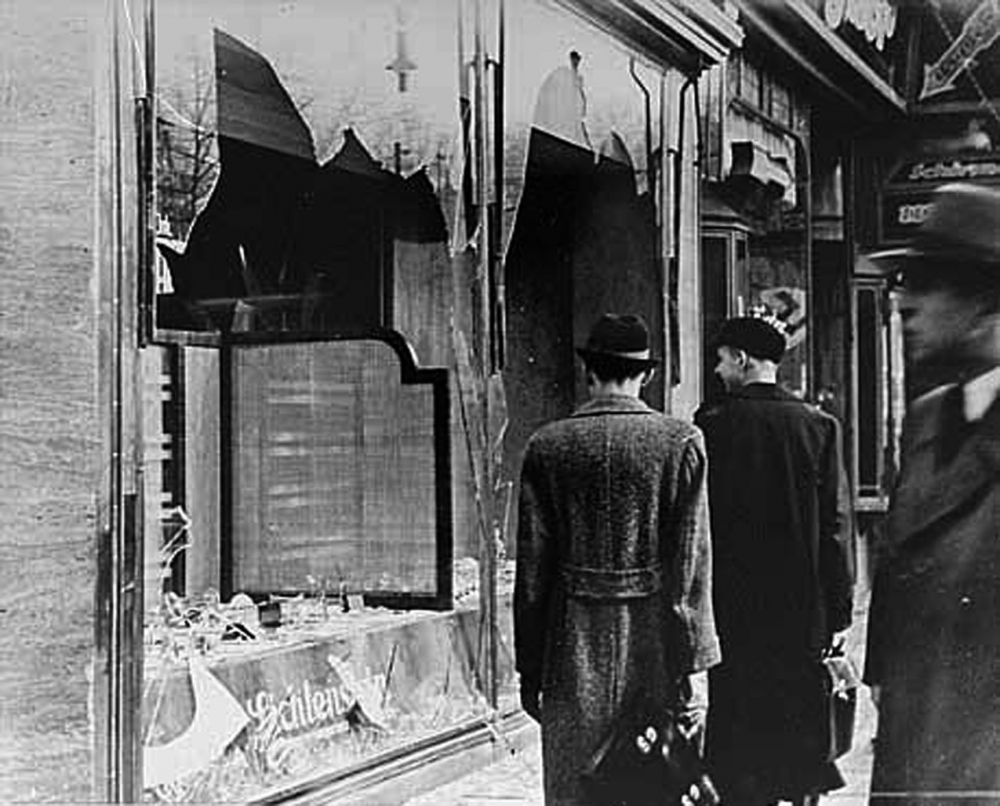 Photo en noir et blanc de trois hommes portant des chapeaux et des manteaux, se tenant sur le trottoir et regardant deux vitrines de magasin endommagées. Deux hommes ont le dos tourné à la caméra.