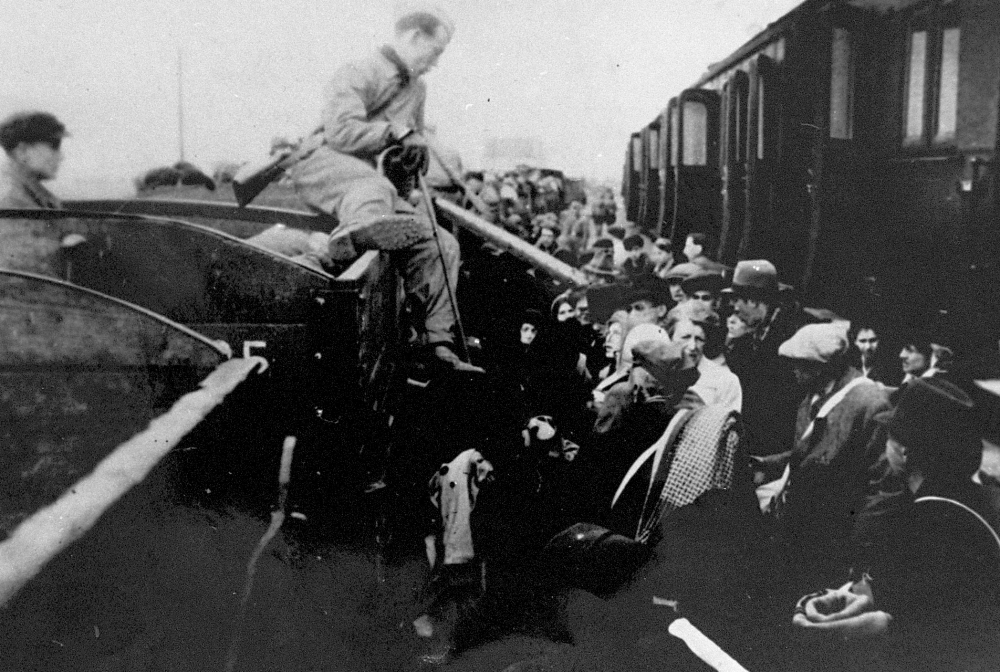 Black-and-white photograph of a man sitting on the edge of a train car, holding a cane, and looking onto a platform full of people.