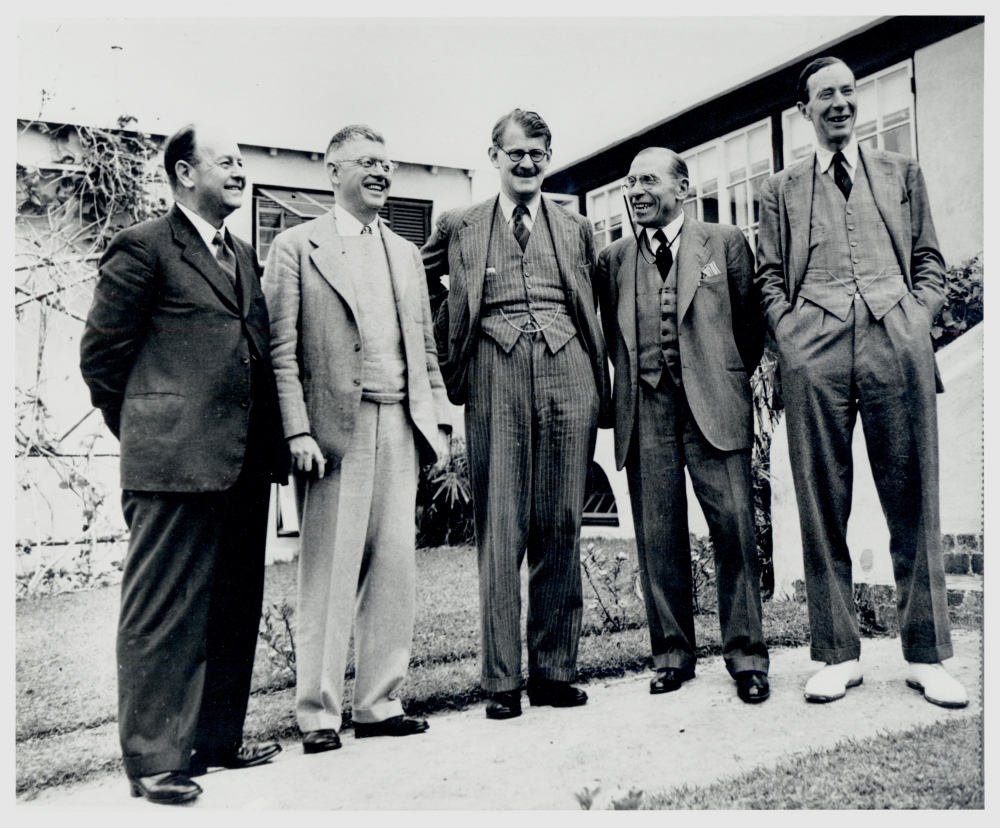 Photo en noir et blanc de cinq hommes, portant des costumes et se tenant en rangée à l'extérieure, souriant et riant.