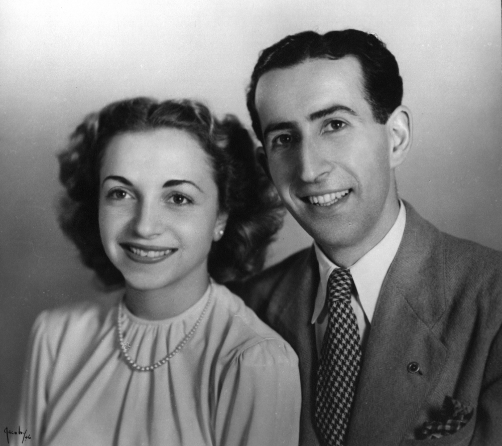 Photo en noir et blanc prise en studio d'un homme et d'une femme assis ensemble et souriant à la caméra. L'homme porte un complet et la femme une blouse avec un collier de perles. Ils ont tous deux des cheveux bruns foncés.