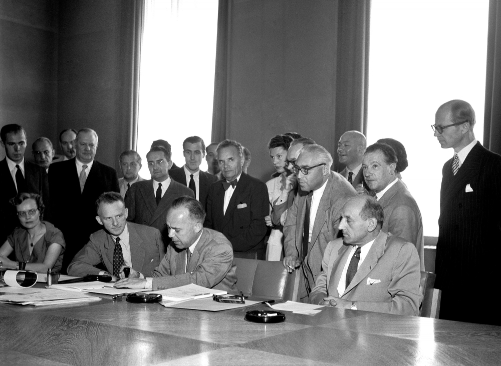 Black-and-white photograph of a group of about 25 people gathered together behind a large table in a conference room. Four men sit at the table, and one man is signing a document. The other people stand behind.