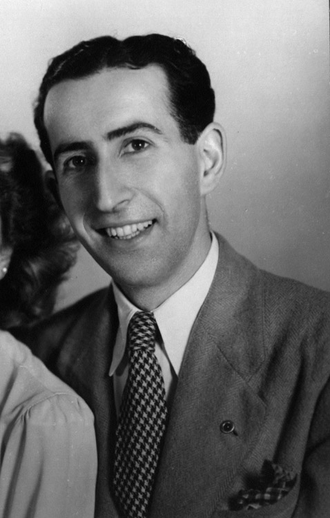 Photo en noir et blanc prise en studio d'un homme assis, souriant à la caméra. Il a des cheveux bruns foncés.”