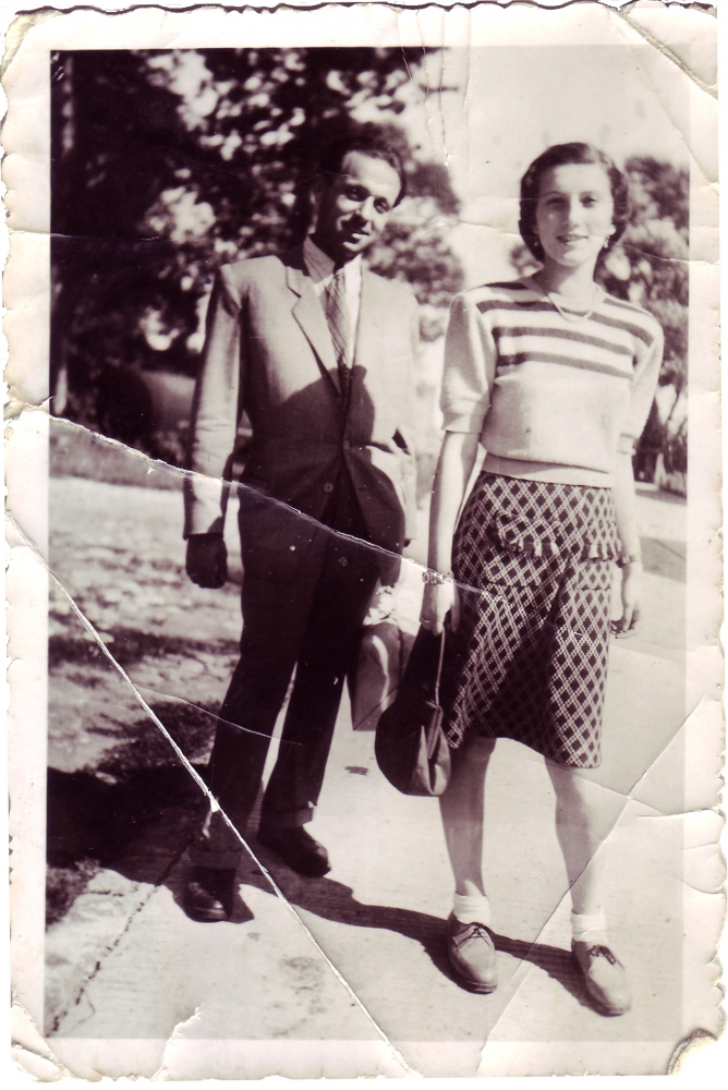 Black-and-white photograph of a man and woman standing together outdoors on the pavement, with trees in the background. The man wears a suit, and the woman wears a blouse and polka-dot skirt.