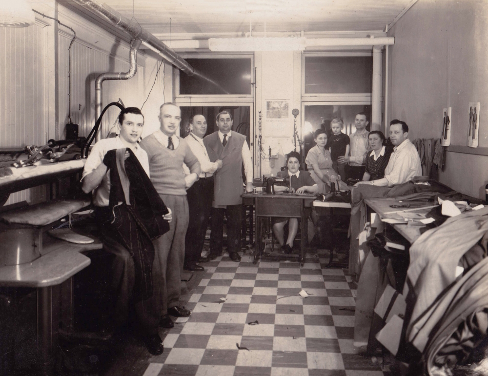 Photo en noir et blanc d'un groupe d'environ dix personnes debout ou assises ensemble dans une salle dans laquelle il y a deux fenêtres au fond. Le groupe regarde la caméra. Il y a des piles de tissus et de vêtements à droite et le plancher est à motif de damiers.