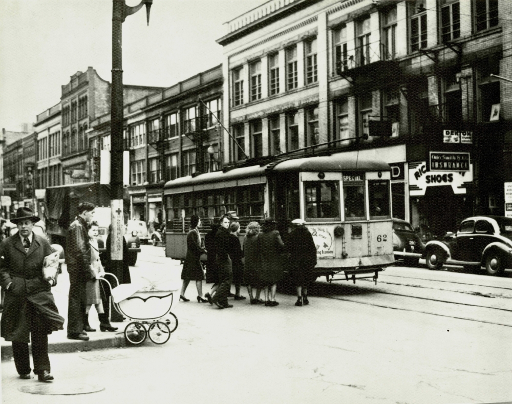 Photo en noir et blanc d'une scène d'une, prise au niveau de la rue. Un groupe de personnes attendent pour embarquer dans un tramway stationné dans la rue.